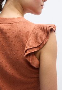 Πλεκτό τοπ σε σκουριά απόχρωση με υφασμένο σχέδιο αστεριού, διαθέτοντας σχεδιασμό με βολάν στους ώμους και ριμπ λαιμόκοψη για επιπλέον λεπτομέρεια.