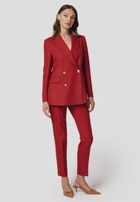 Czerwona marynarka dopasowana, podwójnie zapinana na złote guziki, z dwiema kieszeniami oraz dopasowanymi wąskimi spodniami. Gładki materiał; beżowe szpilki z ostro zakończonym noskiem.