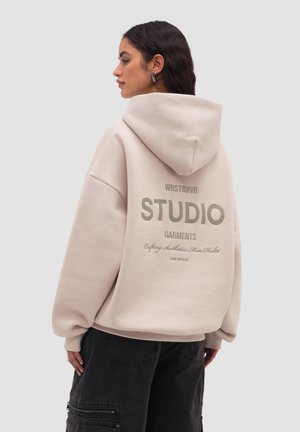 Žena s tmavými vlasy, která má na sobě béžovou volnou mikinu s kapucí s nápisem „WRSTBHVR STUDIO Garments“ na zádech a černé kalhoty s kapsami.