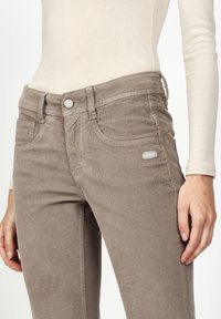 Brown corduroy pants with a snug fit, featuring front pockets, tonal stitching, and a metallic button at the waistband.