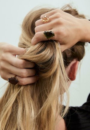 Due an anelli indossati sulle mani di una persona con lunghi capelli biondi. Un anello è rettangolare, di colore marrone chiaro e oro, mentre l'altro è esagonale, di colore verde scuro e nero.
