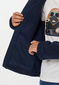Άτομο που φοράει μπλε σκούρο fleece μπουφάν, δείχνοντας την εσωτερική τσέπη και ένα λευκό μπλουζάκι με γραφικό σκύλου DJ.