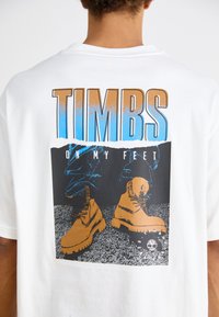 Camiseta blanca con un diseño gráfico que incluye el texto "TIMBS ON MY FEET" y una ilustración de botas marrones sobre un fondo negro.