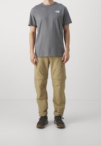 Harmaa puuvillapaita lyhyillä hihoilla ja valkoisella logolla. Vaaleat cargo-housut vetoketjullisilla taskuilla. Mustat vaelluskengät, joissa on tekstuuripohjat.