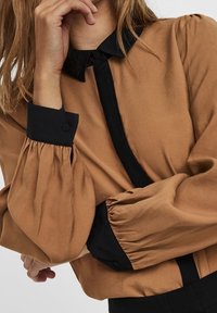 Blusa di raso marrone con colletto, patta e polsini neri; maniche puff con texture arricciata e dettagli in bottone neri.