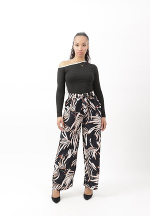 Frau mit langen lockigen Haaren trägt ein schwarzes schulterfreies Oberteil, schwarze weite Hosen mit beigem Blattmuster und schwarze High Heels, steht vor weißem Hintergrund.