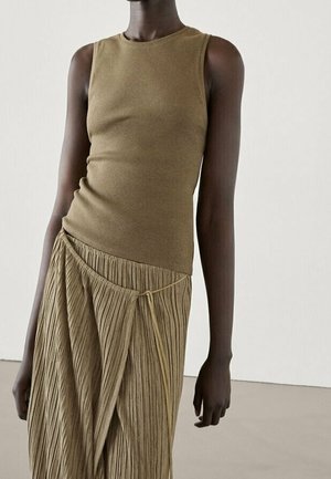 Personne portant un haut sans manches vert olive et une jupe plissée texturée assortie avec un détail de nœud à la taille sur un fond uni.
