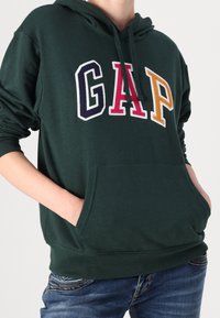 Felpa verde con tasca a marsupio, con logo "GAP" ricamato in blu navy, rosso e giallo. Il materiale è morbido con polsini a coste.