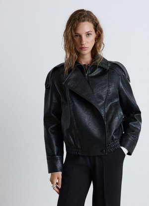 Woman with wavy hair wearing a black textured leather jacket and black pants, standing against a plain light gray background.