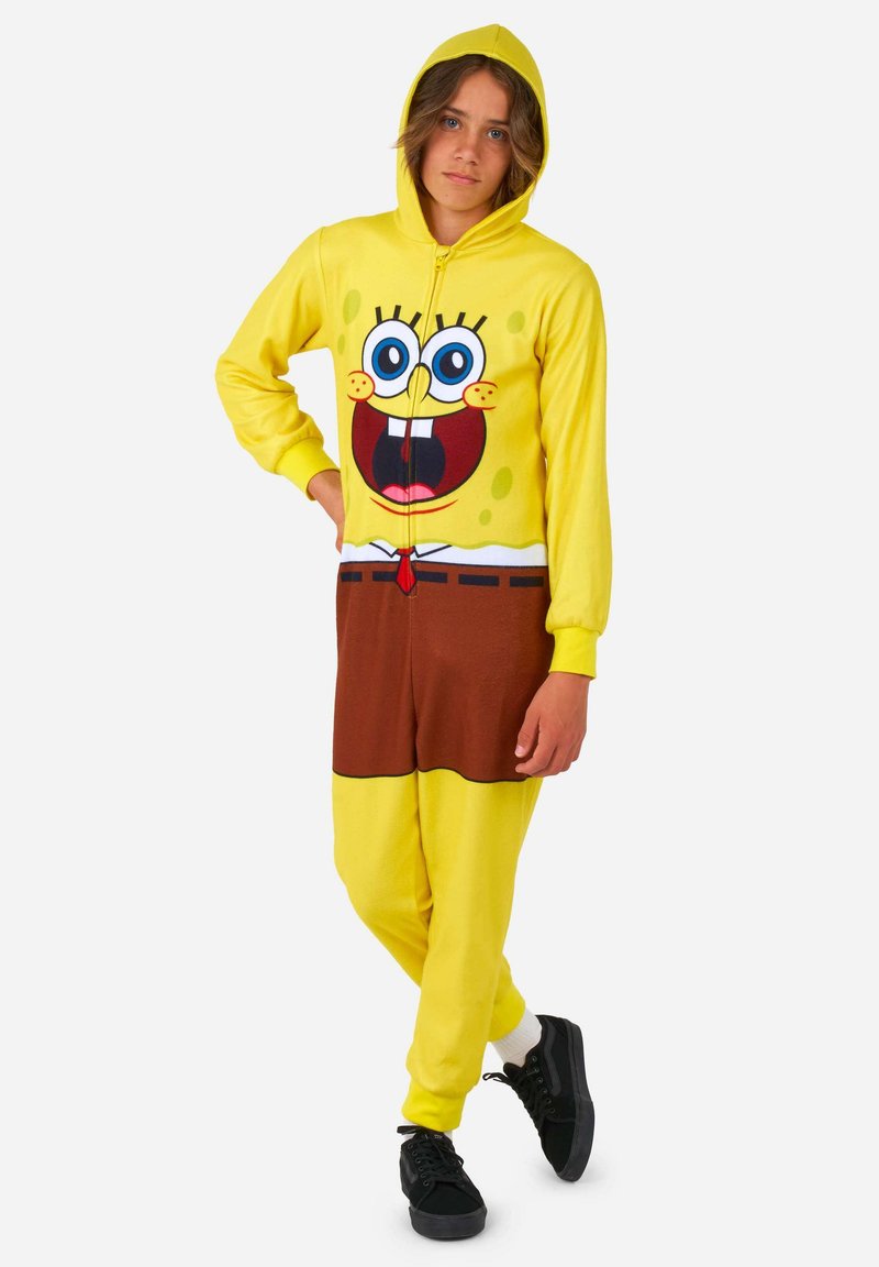 Un mono amarillo con un diseño de personaje de dibujos animados que tiene una cara sonriente, fondo marrón, mangas largas y cierre de cremallera frontal.