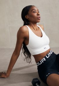 Valge sportlik spordirinnahoidja V-kaeluse disainiga ja logoga, koos mustade kõrge vöökohaga lühikeste pükstega, millel on julge vöökoht ja logodetail.