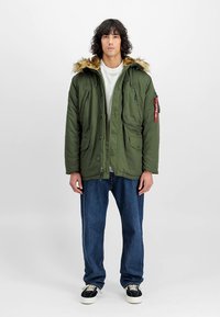 Oliwkowo-zielona parka z kapturem wyściełanym futrem, zapinana na zamek i guziki, z dwoma kieszeniami z przodu oraz czerwoną metką z logo na ramieniu. Nosi się ją na białej koszuli.