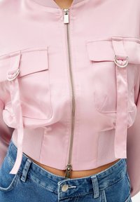 Rosa satin bomberjacka med framside dragkedja, dubbla bröstfickor och ribbad nederkant. Har glänsande detaljer och en slät yta. Bärs tillsammans med denim.