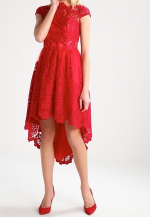 Kvinde iført en rød blonder kjole med højt-lavt snit og bølgede kanter samt røde højhælede sko, stående foran en ensartet hvid baggrund.