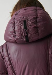 Lila puffjacka med en hög, samlad huva, slät textur och en svart tygetikett med "Clarks" broderat i lila.