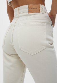 Jeans de mezclilla blanca con una cintura alta, pierna recta y diseño clásico de cinco bolsillos, con una etiqueta de marca de cuero marrón en la parte trasera.