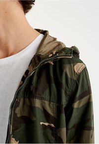 Giacca con motivo camouflage in verde scuro e beige, dotata di chiusura con zip, cappuccio regolabile e tessuto morbido e leggero.