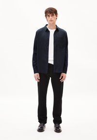 Marineblauw overhemd met knopen en een zak, gecombineerd met een wit T-shirt met ronde hals, zwarte broek en zwarte schoenen in een minimalistische setting.