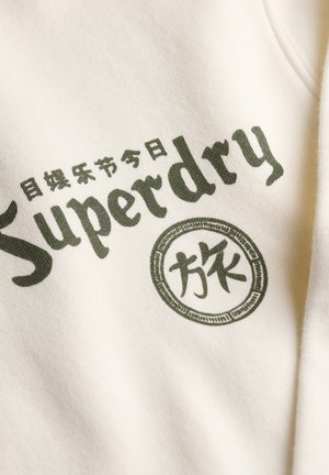 Primo piano di un tessuto crema con il testo "Superdry" in verde scuro e caratteri giapponesi stampati su di esso.