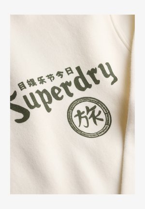 Close-up van een crème stof met donkergroene "Superdry" tekst en Japanse karakters erop gedrukt.