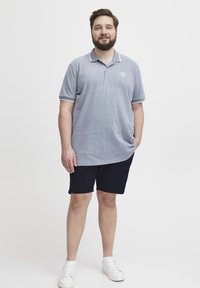 Graues Polohemd mit weißen Akzenten, kurzen Ärmeln und einem Logo auf der Brust, kombiniert mit navyblauen Shorts und weißen Turnschuhen.