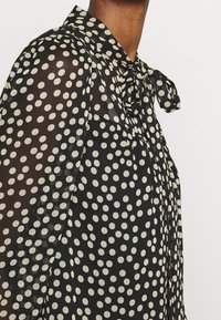 Blusa nera trasparente con grandi pois bianchi, colletto a fiocco e maniche lunghe. Tessuto leggero con una consistenza delicata.