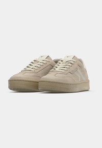 Beige Wildleder-Sneaker mit strukturierten Seiten, cremefarbenen Schnürsenkeln und einer dicken Gummisohle mit Musterprofil. Weiße Logo-Akzente auf der Zunge.