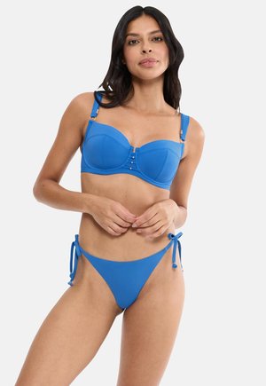 Woman wearing a blue bikini with underwire top and side-tie bottoms, posing with hands clasped at waist against a plain background.