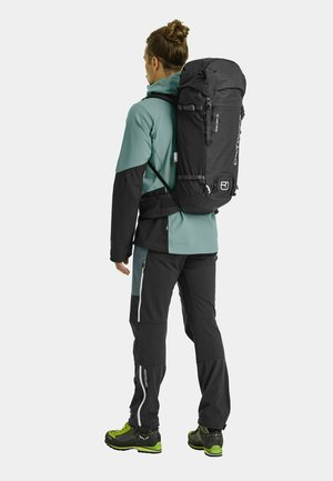 Schwarzer Wanderrucksack mit Roll-Top-Design, getragen über einer türkis- und schwarzfarbenen Jacke. Auffällige Reißverschlüsse an der Hose und grüne Akzente an den Schuhen.
