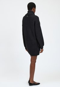 Schwarzer, übergroßer Pullover mit hohem Kragen, strukturiertem Stoff, gerippten Bündchen und Saum. Goldfarbene Akzentdetails auf der Rückseite. Kombiniert mit schwarzen Ballerinas.