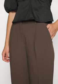 Personne portant un pantalon marron taille haute avec des poches et un chemisier noir à manches bouffantes, une main dans la poche, cadré à la taille et aux cuisses.