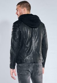 Mann trägt eine schwarze Lederjacke mit angenähter schwarzer Kapuze, kombiniert mit verblassten schwarzen Jeans, von hinten vor einem schlichten hellen Hintergrund gezeigt.