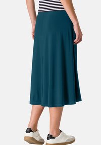 Jupe mi-longue bleu sarcelle en tissu doux. Modèle évasé avec une texture lisse. Portée avec un haut rayé et des baskets blanches.