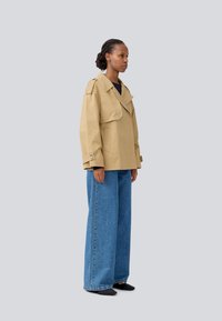 Béžová oversize bunda so širokými reverzami a veľkými vreckami, kombinovaná s voľnými modrými denimovými džínsami a čiernymi plochými topánkami, zobrazená zo strany.
