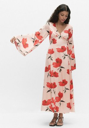 Vrouw draagt een lange, lichtroze jurk met grote rode bloemenprints en brede klokkende mouwen, gecombineerd met zwarte sandalen met bandjes.