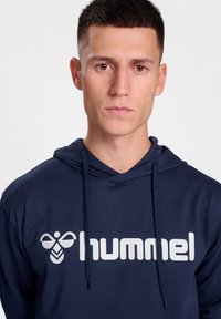 Mörkblå hoodie i mjukt tyg, med en framficka och kontrasterande vit "hummel"-logotyp. Innehåller snörning och en avslappnad passform.