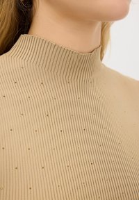 Maglione beige a collo alto a coste con un motivo testurizzato, caratterizzato da piccole screziature dorate. Materiale morbido e design aderente.