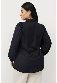 Donkerblauwe bloes met opbollende lange mouwen, een kraag en een gladde textuur. Van achteren weergegeven met een gebogen zoom en met knopen sluitende manchetten.