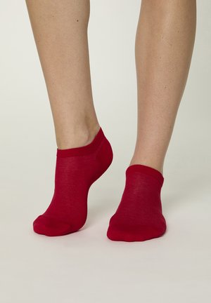 Füße mit leuchtend roten Knöchelsocken, ein Fuß flach auf dem Boden, der andere auf den Zehen erhoben vor neutralem Hintergrund.