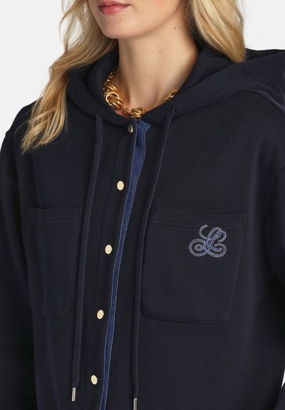 Nő sötétkék kapucnis pulóverben, elülső zsebekkel, arany gombokkal és aranylánc nyaklánccal.