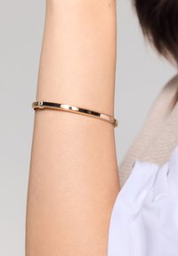 Złota metalowa bransoletka w postaci gładkiego i smukłego wzoru, z zapięciem i delikatną fakturą, spoczywająca na nagim ramieniu.