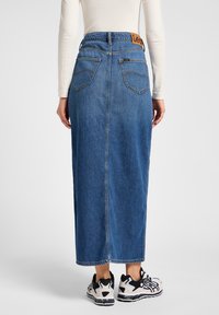 Denim maxi skirt in a solid blue hue, featuring a high waist, two back pockets, and a tan leather brand patch. Includes a straight cut.