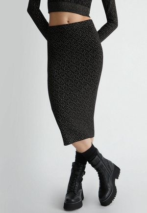 Rajattu näkymä henkilöstä, joka pukeutunut mustaan kuvioituun pitkähihaiseen lyhytpaitaiseen yläosaan, siihen sopivaan midihameeseen ja mustiin paksukorkoisiin maiharikenkiin.