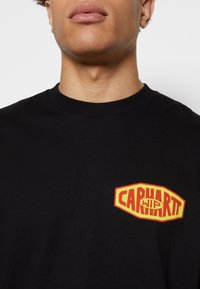 Muž v černém tričku s kulatým výstřihem, na němž je vlevo na hrudi viditelné logo "CARHARTT WIP" v červené a žluté barvě, viditelné od ramen po horní část hrudi.