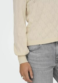 Suéter de punto color crema con un patrón de diamantes texturizado, puños acanalados y dobladillo. Combinado con jeans de mezclilla gris claro.