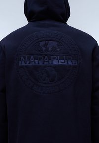 Felpa navy con cappuccio caratterizzata da un grande logo ricamato testurizzato sul retro, con elementi di design circolari e dettagli di branding prominenti.