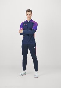 Marinefarbener Sportanzug mit lila Farbverlauf-Ärmeln, ausgestattet mit einem hohen Kragen und rosa Akzenten. Enthält Logodetails auf dem Shirt und der Hose.