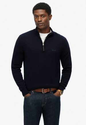 Mørkeblå striktrøje med lynlåskrave, parret med en hvid t-shirt og mørke jeans. Indeholder et læderbælte og et guld-toneur.