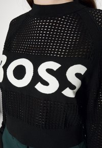 Svart stickad tröja med öppen nätdesign och solid svarta sektioner. Har en stor vit "BOSS"-logotyp på framsidan och ribbad halsringning.