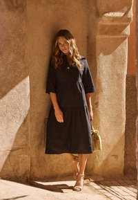 Svart knälång klänning med flikkrage, trekvartsärmar, ögla detaljer längs nederkanten, matchad med guldklacksandaler och en ljus väska.